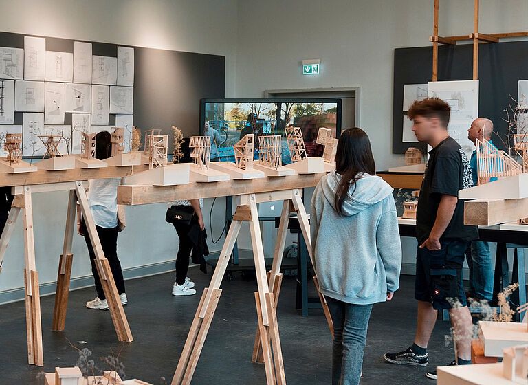 Ein Eindruck aus der Ausstellung zur Werkschau der Fachrichtung Architektur. Zu sehen sind Besucher, die sich Modelle und Zeichnungen ein einem Seminarraum anasehen.