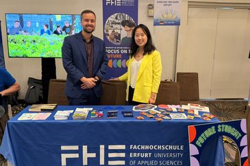 Kevin Rausch und Ha Giang Nguyen vom International Office der Fachhochschule Erfurt pr&auml;sentieren den Messestand der Hochschule mit Informationsmaterialien und Werbeartikeln. Im Hintergrund sind Roll-ups mit dem Logo der FHE sowie ein Bildschirm mit Einblicken in das Hochschulleben zu sehen.