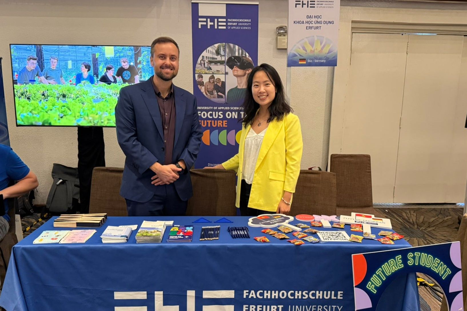 EHEW-Messe Ho Chi Minh City Kevin Rausch und Ha Giang Nguyen vom International Office der Fachhochschule Erfurt präsentieren den Messestand der Hochschule mit Informationsmaterialien und Werbeartikeln. Im Hintergrund sind Roll-ups mit dem Logo der FHE sowie ein Bildschirm mit Einblicken in das Hochschulleben zu sehen.