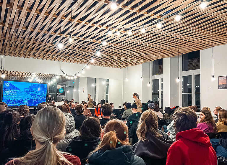 Foto einer Wir-Müssen-Reden Veranstaltung. Im Vordergrund sieht man das mit dem Rücken zugewandte Publikum, Im Hintergund ist links ein großer Monitor, rechts daneben die Gäst:innen und Veranstalter:innen.