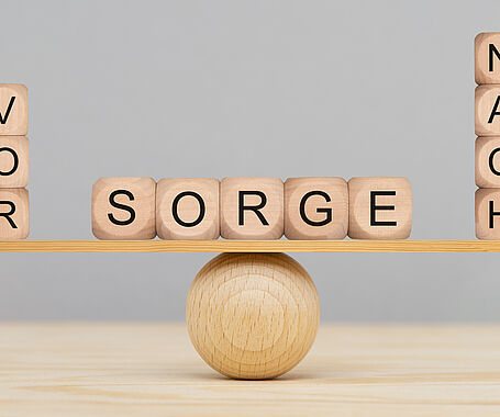 Darstellung einer ausbalancierten Wippe mit drei Holzwürfeln auf der linken Seiten, dies das Wort Vor ergeben, vier Holzwürfeln auf der rechten Seite, die das Wort Nach ergeben und in der Mitte fünf Würfeln, dies das Wort Sorge ergeben. Vorsorge ist besser als Nachsorge