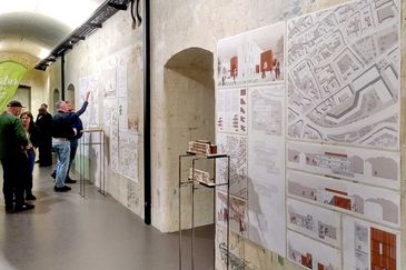 Schr&auml;ger Blick entlang einer verwitterten Gew&ouml;lbewand mit Entwurfsplakaten zum Naturkundemuseum Erfurt: Stadtpl&auml;ne, Schnitte, Grundrisse und eine Visualisierung mit rotbraunen Fassadenelementen. Davor stehen zwei Modelle auf Metallst&auml;ndern, im Hintergrund unterhalten sich mehrere Besucherinnen und Besucher.