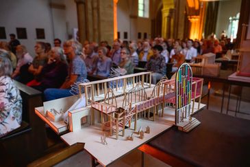 Im Vordergrund steht ein helles Architekturmodell aus Holz mit filigranem Ger&uuml;st und einem bunten Glasfenster. Dahinter sitzt unscharf das Publikum in den Bankreihen einer Kirche und schaut nach vorn.