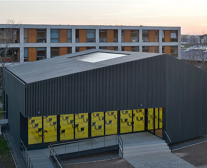 Im Bild: Der Green Campus von schräge oben fotografiert. Im inneren ist das Licht eingeschaltet, im Hintergrund sieht man den hellblau-pinken Sonnenuntergangshimmel