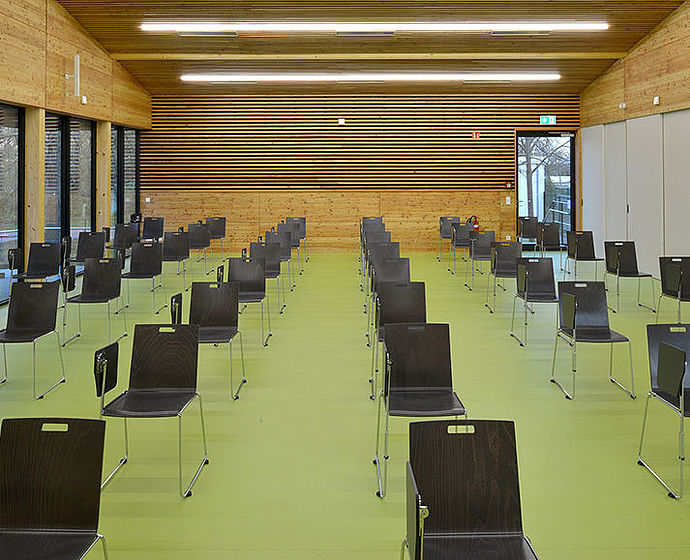 Blick von vorn in den Hörsaal. Der Fußboden ist grün, Wände und Decke aus natürlichem Holz. Im Raum stehen 6 Reihen schwarzer Stühle.
